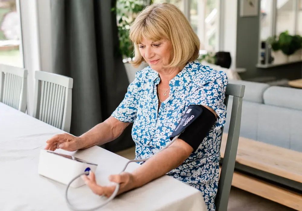 Kvinna sitter vid ett bord och mäter blodtrycket hemma med manschetten runt överarmen